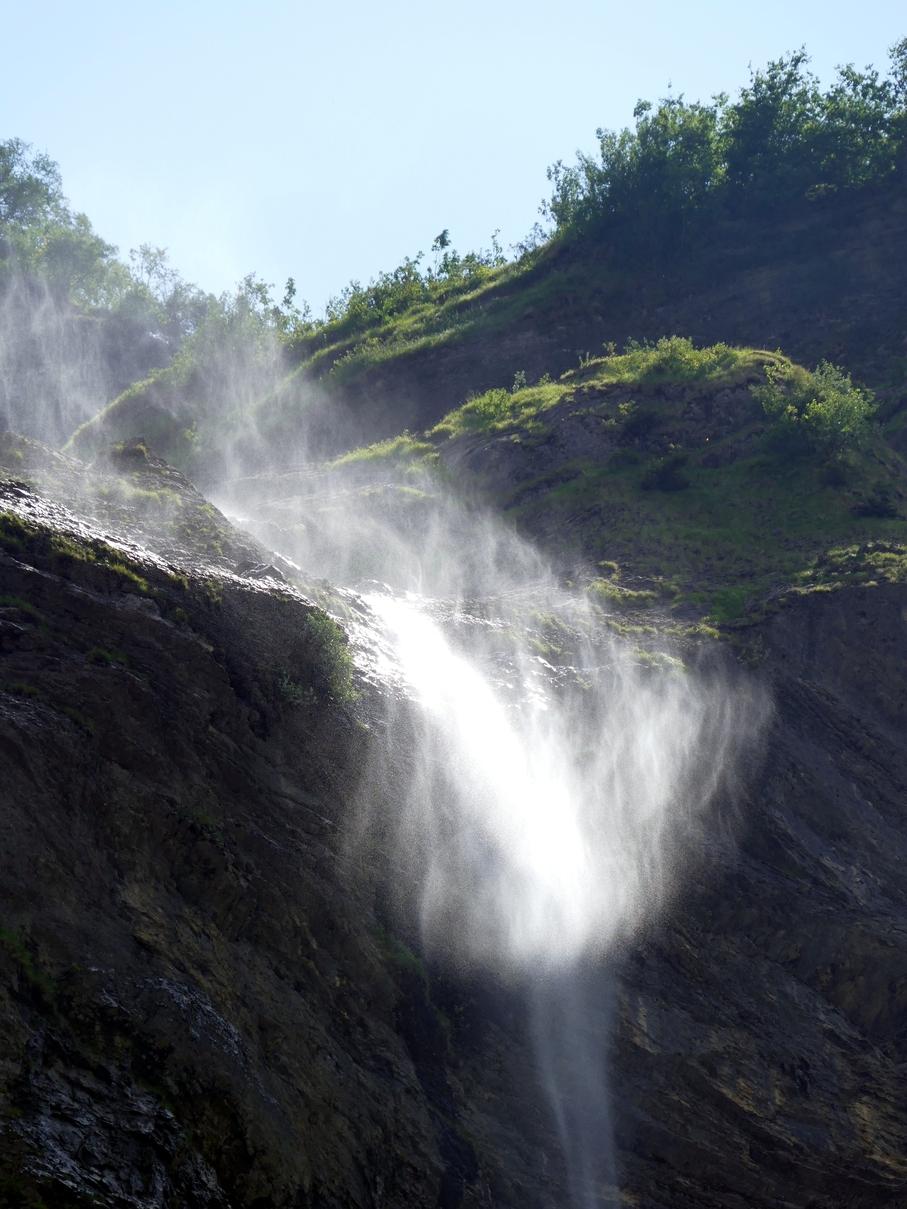Le haut d'une cascade, à l'endroit où elle plonge depuis un rebord herbu. La chute est dans l'ombre, le haut de la cascade a l'air de partir en fumée à cause du vent qui disperse l'eau.